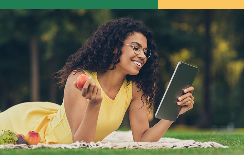 Woman holding an apple and reading a tablet on the grass