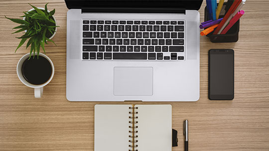 Wooden desk from above including laptop, notebook, plant, phone, pens and markers