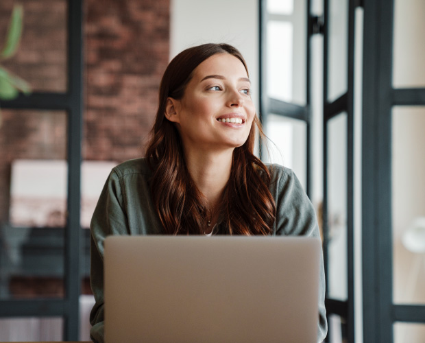 woman sitting by a brightly lit window, looking out, smiling, while working at a laptop