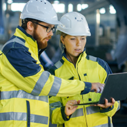 Two workers wearing hard hats and safety jackets sharing a laptop