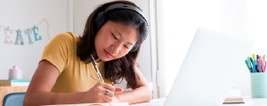 Young student writing in notebook while studying on laptop at desk