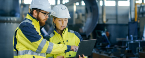 Two workers wearing hard hats and safety jackets sharing a laptop