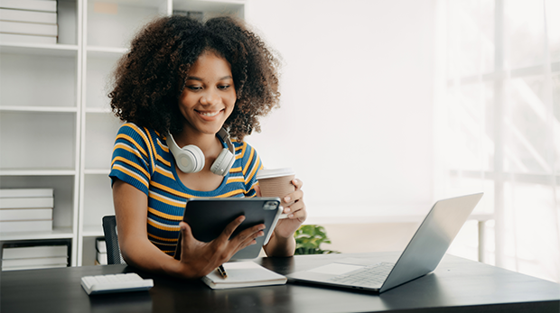 Jonge vrouw zittend aan een bureau kijkend naar een tablet terwijl ze koffie drinkt