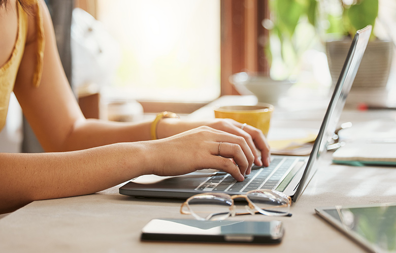 Females hands typing on a laptop with glasses, a tablet and yellow mug on desk with window in the background