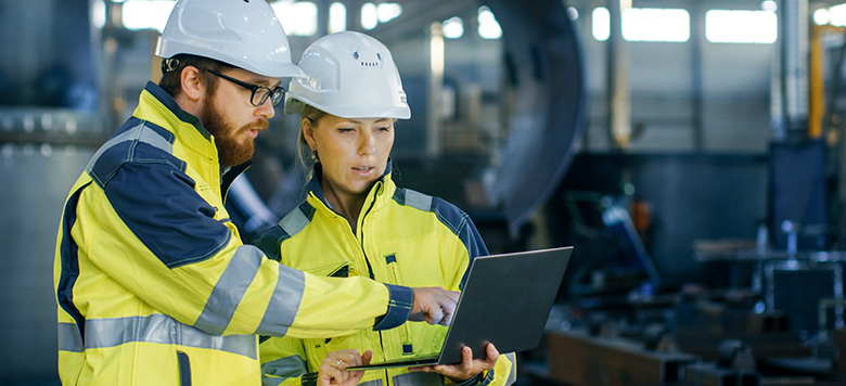 Two workers wearing hard hats and safety jackets sharing a laptop