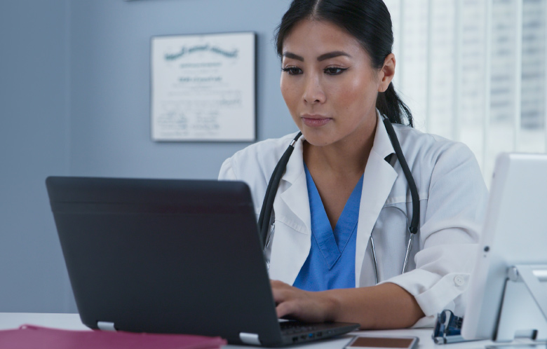 Asian physician sitting at a desk while researching on a laptop.