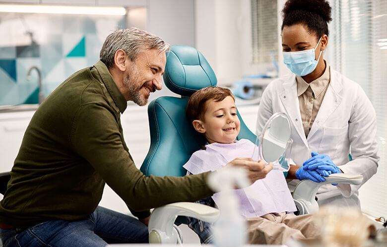 Young boy looking at his teeth in the mirror while sitting in a dentist chair with man and dentist wearing mask standing next to him